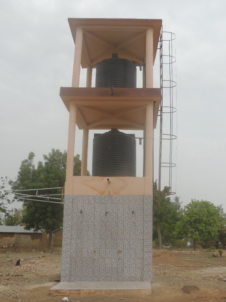2 - Wasser für Afrika - Wasser für Afrika Two-tier outdoor water storage tower with two stacked black tanks, side ladder, on a tiled concrete base in a dry landscape