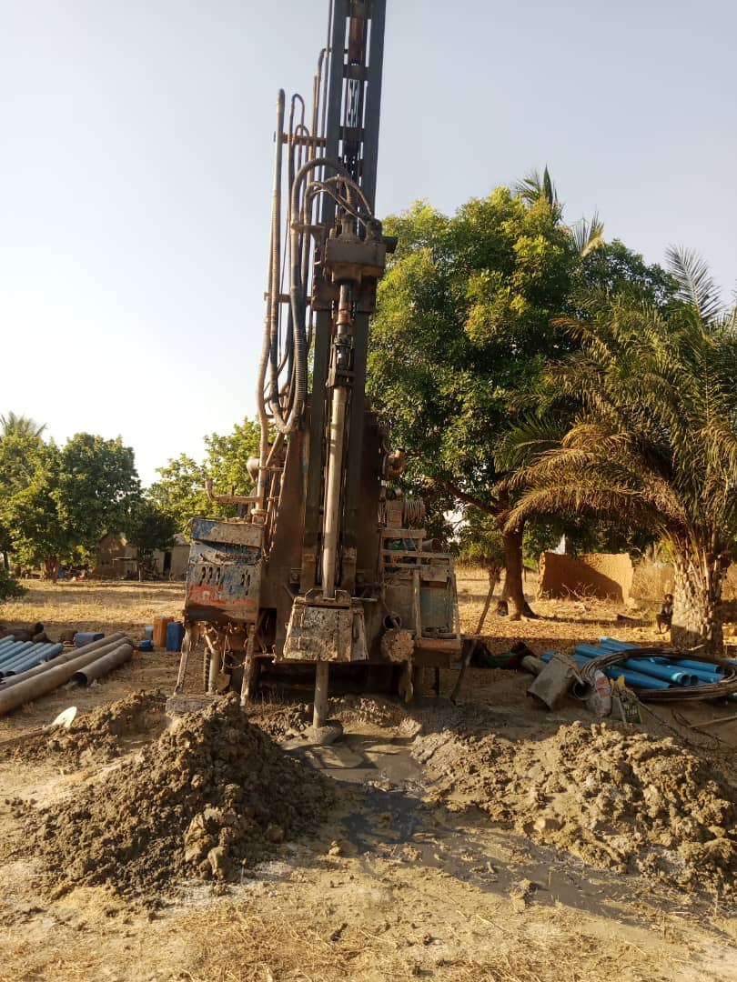 IMG-20251228-WA0023 - Wasser für Afrika - Wasser für Afrika Heavy drilling rig on a dirt construction site with a muddy pile in the foreground and blue pipes nearby.