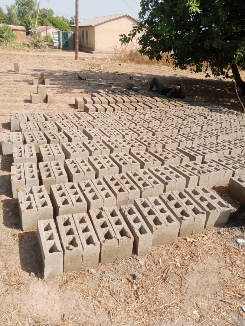 IMG-20260102-WA0017 - Wasser für Afrika - Wasser für Afrika Rows of hollow concrete blocks laid out on a dirt lot for construction, with a person resting under a tree in the background.