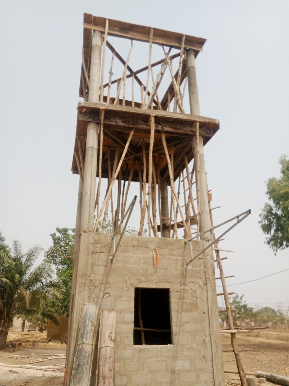 IMG-20260117-WA0005 - Wasser für Afrika - Wasser für Afrika Construction site: a tall wooden scaffolding tower built over a small concrete block building under construction, with planks and braces around it.